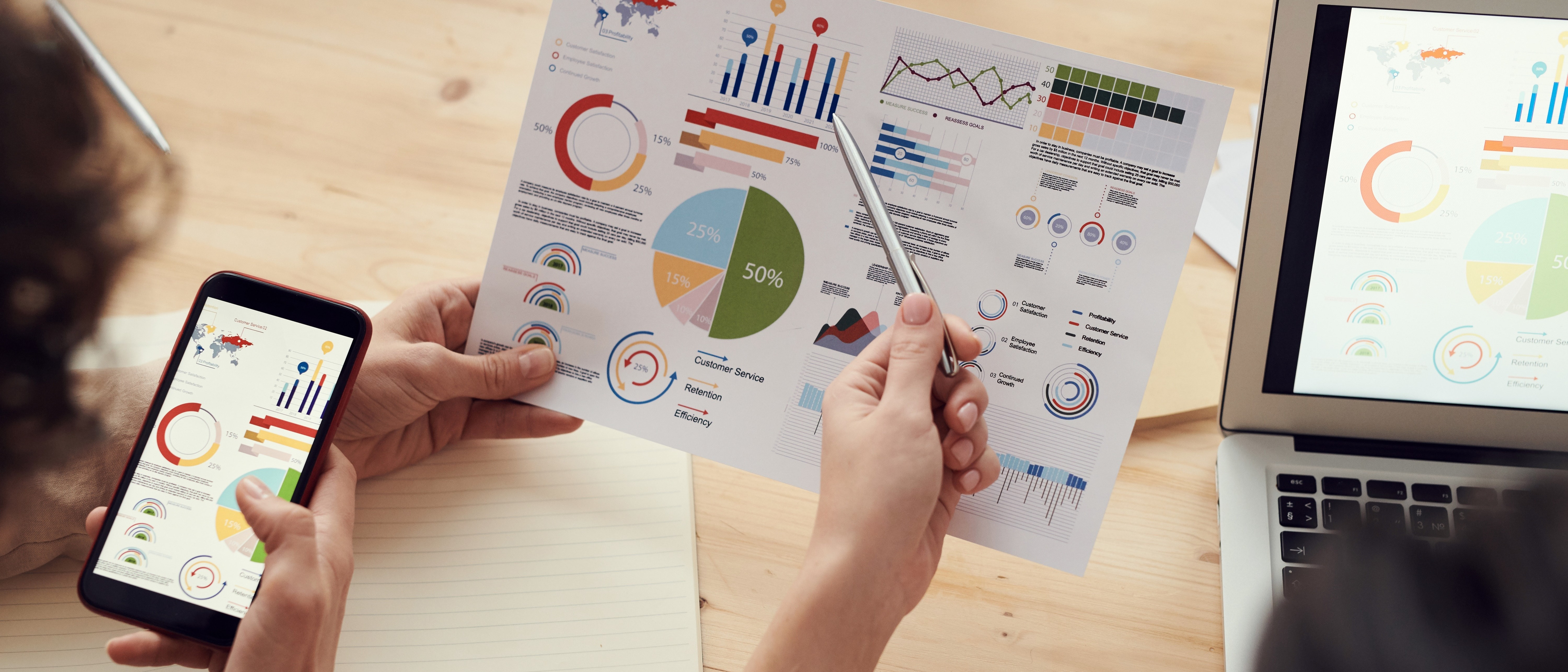 Hands review printed business reports with colorful charts and graphs beside a smartphone and laptop.